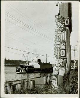 Close-up of a totem pole overlooking a wharf and the vessel Camosun