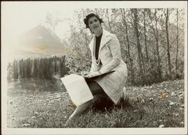 M.P. Iona Campagnolo sits in grassy meadow in front of a forest with mountains in background, reviewing notes, ca. 1975