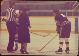 Minister Iona Campagnolo dropping the puck between two unidentified hockey players and an unidentified referee