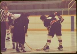 Minister Iona Campagnolo dropping the puck between two unidentified hockey players and an unidentified referee