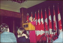Iona Campagnolo gestures from behind a podium labeled “Four Seasons Hotel” in front of a series of draped Canadian flags