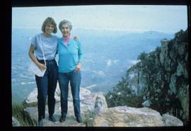 Iona Campagnolo and unidentified woman pose on a cliff above an unknown community overseas