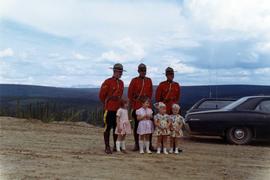Three RCMP constables in dress uniform standing with four small girls in dresses