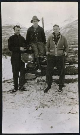 One man sitting on a log in between two men who are standing
