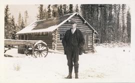 Man standing in front of a log cabin with wagon beside