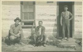Two men sitting beside a house and another standing at the front door