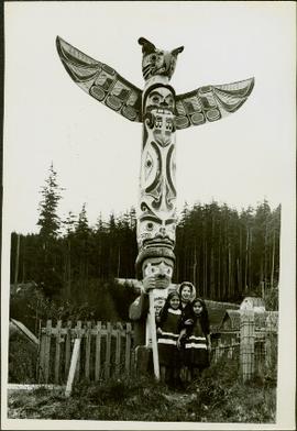 Three young, First Nations girls stand in front of a Memorial Pole
