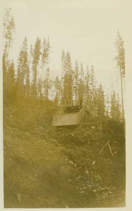 Unidentified man standing at the top of an empty wooden flume (?) situated at the top of a mounta...