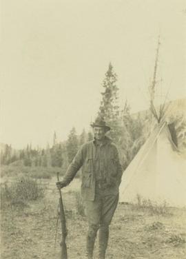 Prentiss Gray standing with rifle in hand in front of a teepee
