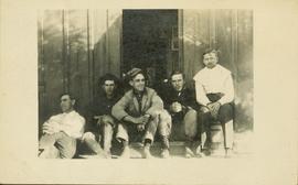 Six men sitting outside the door of a wooden building