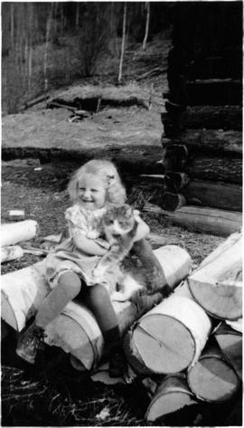 Young Mabel Scholander seated on a wood pile holding a cat