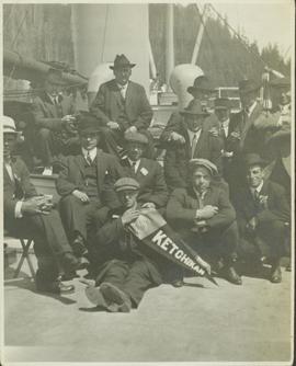 Group photo of men in suits aboard a steamship holding up a "Ketchikan" pennant