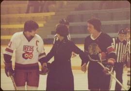Minister Iona Campagnolo standing on ice, holding the hands of two opposing hockey players, one wearing a Chicago Blackhawks jersey
