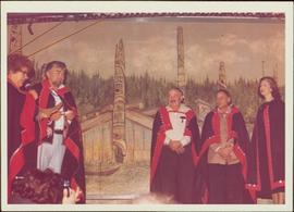 Chief Skidegate Dempsey Collinson, Minister Iona Campagnolo, and three unidentified individuals stand on a stage in button blankets; mural of longhouses and totem poles in background, Haida Gwaii, 1978