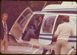 Iona Campagnolo sitting in copilot seat and two unidentified men standing beside helicopter with a “Queen Charlotte” emblem on the side