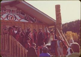 Skeena Riding tour - Group shot of unidentified men and women wearing button blankets in front of longhouse and newly raised totem pole, Kispiox, BC