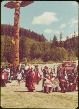 Skeena Riding tour - Distance shot of totem pole flanked by unidentified men, women, and children, some in button blankets