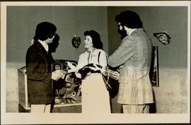 Minister Iona Campagnolo holds a fur craft while speaking with two unidentified men in the Arts and Crafts Division of an unidentified institution