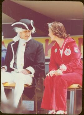 Iona Campagnolo sits on chair in a jogging suit, speaking to a man in George Vancouver costume
