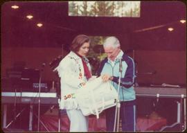 Commonwealth Games, Edmonton 1978 - Iona Campagnolo holds a white leather Canadian Commonwealth Games Team bag with an unidentified gentleman in front of a microphone