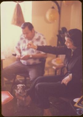 Unidentified man seated beside Iona Campagnolo, who holds a treat above a waiting dog in unknown living room