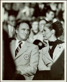 Canada Winter Games, Brandon, MB - Prime Minister Pierre Trudeau and Iona Campagnolo sit in navy and pale blue uniforms in crowded auditorium or arena at the opening of the games