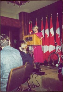 Iona Campagnolo speaks at a podium labeled “Four Seasons Hotel” in front of a series of draped Canadian flags
