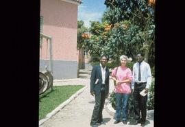 Iona Campagnolo standing between two unidentified men under a tree outside of a school at overseas location