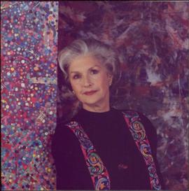 Portrait of Iona Campagnolo in black sweater and multicoloured vest, leaning against a paint-splattered post