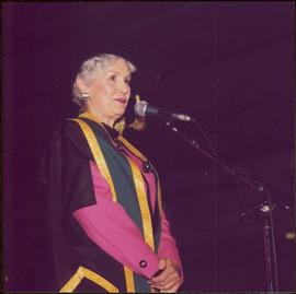 Iona Campagnolo speaking at a microphone in Chancellor’s regalia