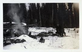 Man on snowshoes crouching in front a fire next to a shack