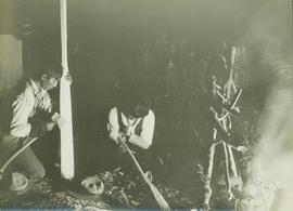 Two unidentified First Nations men making wooden paddles by an evening camp fire