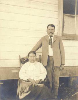 First Nations man standing next to a First Nations woman seated in a rocking chair