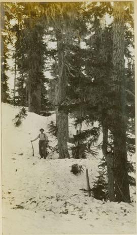 A man, leaning on a rifle, standing on the snow-covered ground next to a tree