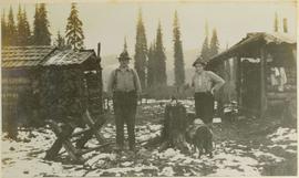 Two men, a cat, and a dog wearing a pack standing between two log cabins