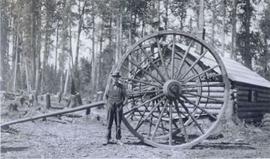 Men posing by large wagon wheels and axle for hauling logs with log cabin behind