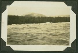 Water view of the Ne Parle Pas Rapids, Peace River; treed shoreline and mountain visible in backg...