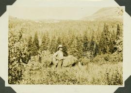 Al Phipps standing beside a horse, tall grass stands before them and a forested landscape and mountain range visible in background