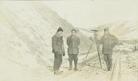 Three men stand with a leveling instrument in the middle of a set of railway tracks in the winter