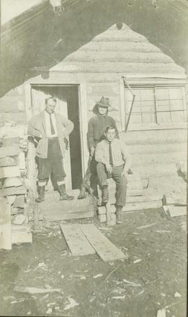 Three men posing outside of a log building