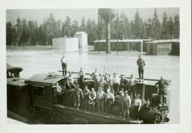 Group photo of twenty-one men standing on top of a railcar situated within a flooded CNR yard at Pacific Station